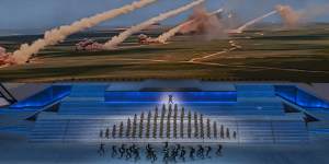 Performers dressed as the military perform in front of a screen depicting firing rockets. 