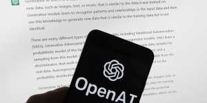 The OpenAI logo is seen on a mobile phone in front of a computer screen displaying output from ChatGPT.