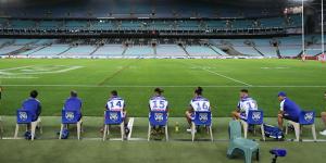 Bulldogs interchange players and support staff,seated well apart,look on at an empty ANZ Stadium.