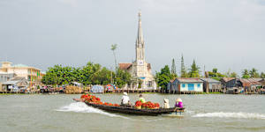 Traditional wooden boats cross the Mekong with Cai Be church in the background.