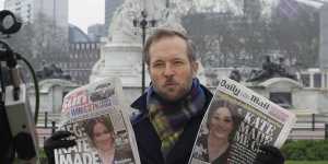 A television journalist holds up two British newspapers as he speaks to camera outside Buckingham Palace.