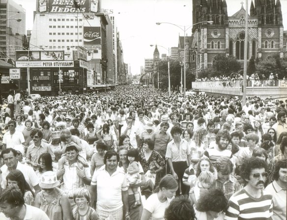 The crowd follows behind the Moomba parade, walking along Swanston Street towards Alexandra Gardens. Photograph taken on Princess Bridge looking up Swanston Street. Flinders Street Station is on the left and St Paul's Cathedral on the right, 1979.