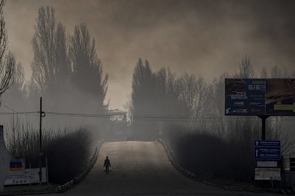 A man carries shopping bags as heavy smoke from a warehouse destroyed by Russian bombardment casts shadows on the road outside Kyiv, Ukraine.