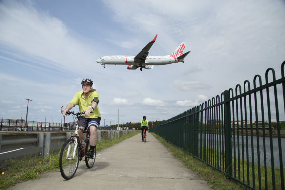 A Virgin Australia Boeing 737 flies over cyclists on the Alexandra Canal Cycleway near Sydney Airport.
