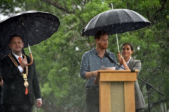 Prince Harry speaks to the community as his wife Meghan, the Duchess of Sussex holds an umbrella under heavy rain.