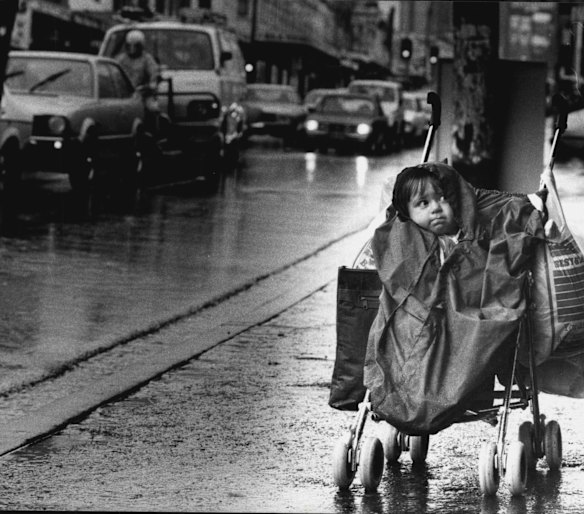 Parramatta Rd in 1984. Kristy Jackson of Leichhardt  protected from the rain and cold as she waited for her mother Lynda Jackson. 