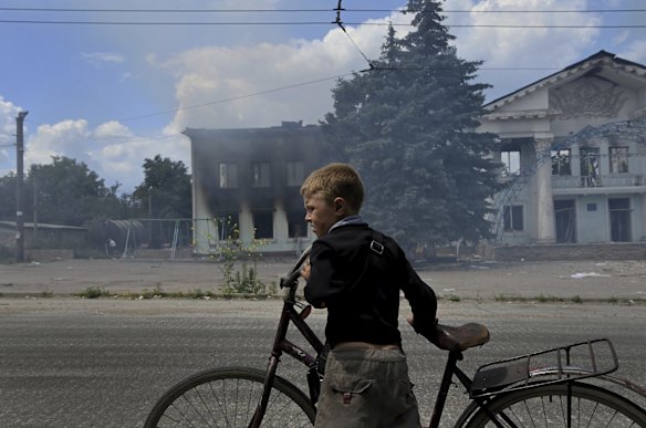 Timosha 8yrs stands with his bicycle in-front of the still burning Palace of Culture in Lysychansk that was destroyed by an air-strike that killed four people the day before. Lysychansk, Ukraine. 
