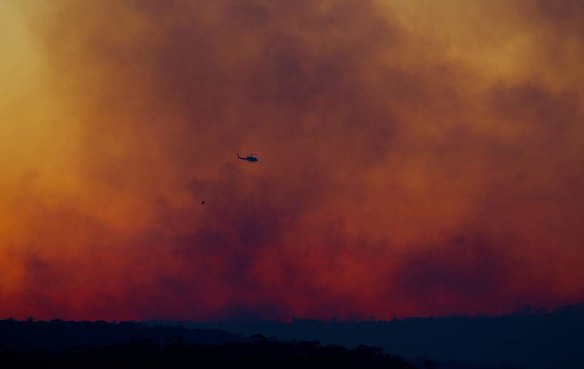 A water bombing helicopter flies through plumes of smoke from the State Mine Fire at dusk along the Bells Line of Road in the Blue Mountains.