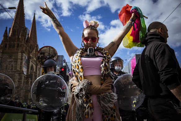 A colourful protester with a camera strapped to her chest is backgrounded by police officers in riot gear.