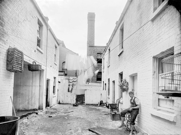 Location unknown, c1901. The location of this whitewashed courtyard is not known but the photograph was taken around 1901. A young barefoot man sits on a sewing table. Wicker baskets are hung up above the door opposite, there's a washing copper nearby, and laundry drying on the line. This scene of domestic intimacy is in contrast to the industrial chimney looming nearby.