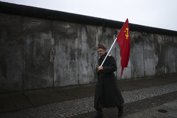 An elderly man with a Soviet flag walks in front of remains of the Berlin Wall.