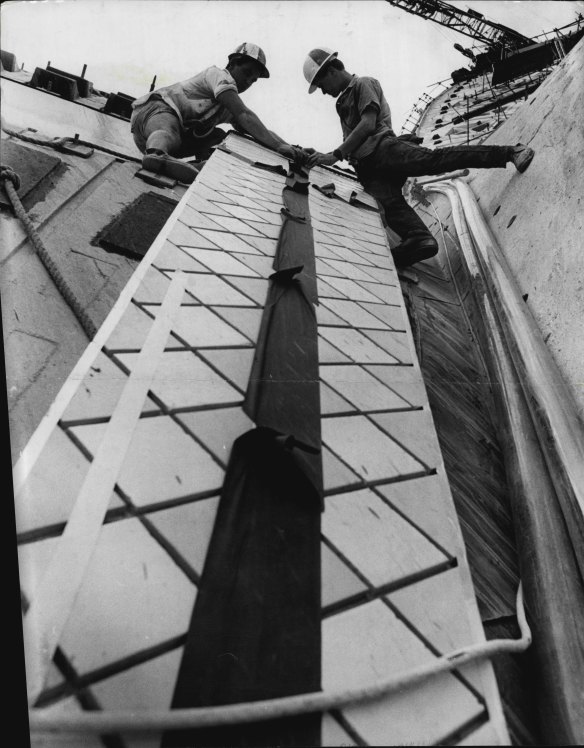 “Mountaineers” of the Opera House, workmen Phil Manion, of North Sydney (left), and David Bain, of Darling Point, perched high on a vertical sail roof as they inspect the first test strip of tiling in January 1965.