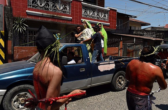 Two self flagellating masked men walk past a group of young men standing around a religious statue on the back of a ute.