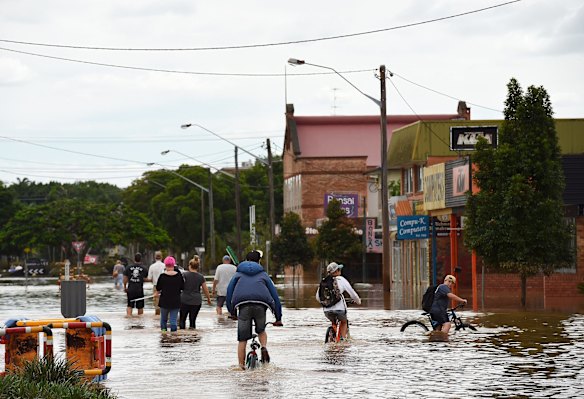 Teenagers cycle behind a group of people with brooms wading through the water of the flooded streets of Lismore.