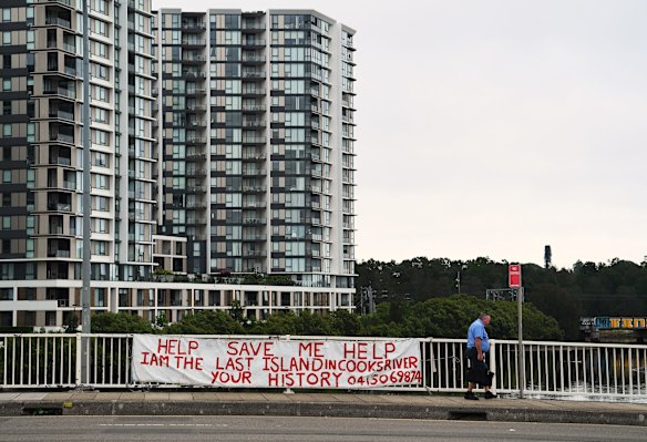A man walks over the Cooks River on a foot bridge, past a sign about saving the last island on the Cooks River which is called Fatima Island , Wolli Creek, Sydney.