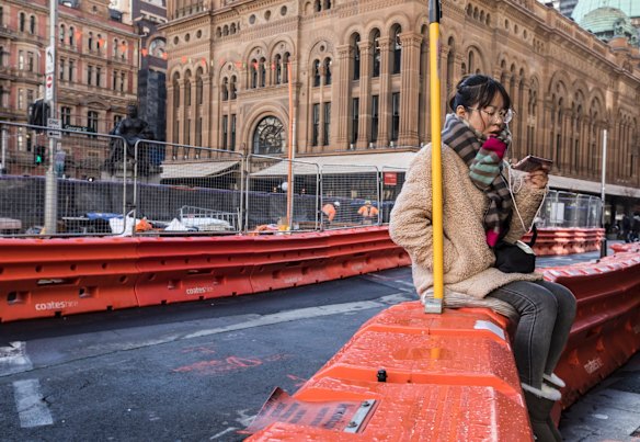 A woman sits transfixed by her phone by a sign advertising Ugg Boots amongst the chaos of the construction of the light rail at the intersection of George Street and Bridge Street, Sydney.