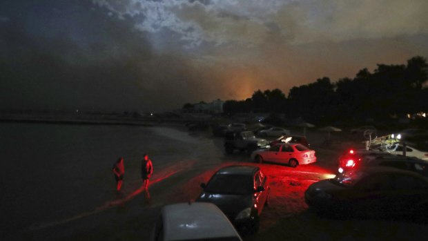 People stand in the sea near to cars parked on the beach to protect them from the fire in the town of Mati.