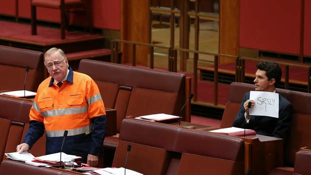 Liberal Senator Ian Macdonald speaks on the mining tax repeal while Scott Ludlam holds up a sign. Photo: Alex Ellinghausen