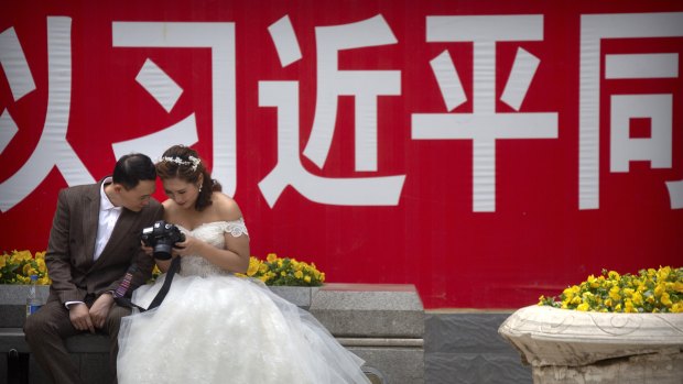 A bride and groom look at photos on the back of a camera near a propaganda slogan with the name of Chinese President Xi Jinping in Beijing.