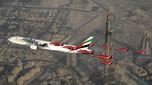 A formation of nine Royal Air Force Red Arrows Hawk aircraft fly alongside a Boeing 777 aircraft at the start of the Dubai Airshow.