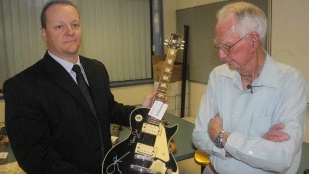 Detective Senior Constable Lonie Horn holds up one of the recovered guitars as its owner, Leonard "Merv" French, looks on.