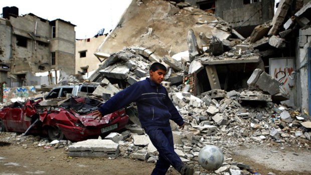 A Palestinian boy plays football in front of a destroyed house.