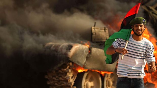 Rebels advance ... a man holds the Kingdom of Libya flag as he walks past a burning wrecked tank in Ajdabiya.