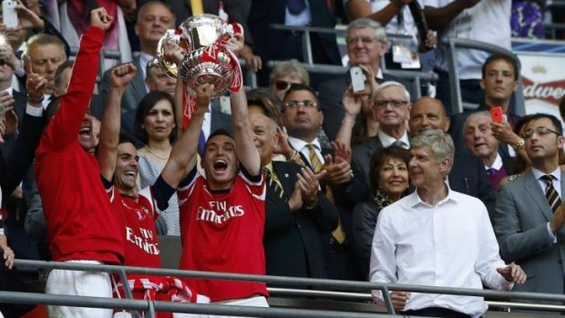 Wenger watches on as Thomas Vermaelen and Mikel Arteta hold up the trophy.