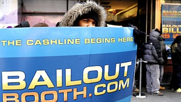 A worker displays a sign advertising a free money handout and a website, as people line up to receive cash from Bailout Bill at the Bailout Booth on Times Square in New York.