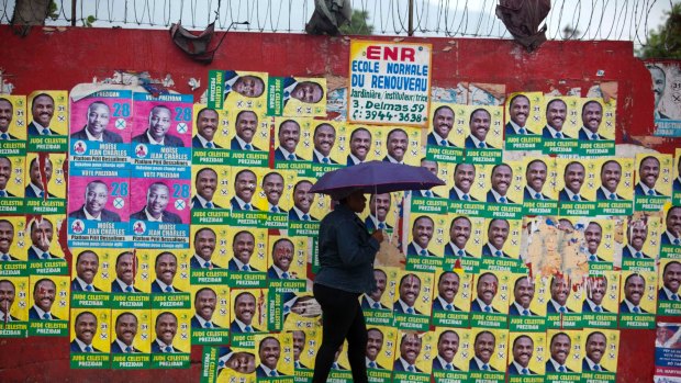 A woman walks with an umbrella as protection from a light rain, backdropped by a wall blanketed with presidential campaign posters, in Port-au-Prince, Haiti.