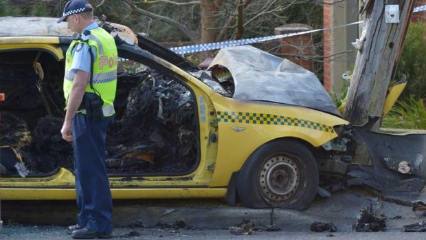 A police officer examines the burnt-out wreckage of a taxi in Glen Waverley yesterday. The taxi's driver had been stabbed to death by a passenger, who left his victim lying on the road before stealing the cab and then being incinerated after crashing into a power pole.