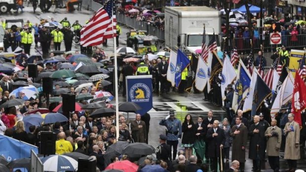 Survivors, families of victims, officials, first responders and guests pause as the flag is raised at the finish line of the Boston Marathon.