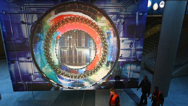Workers walk past a giant photograph of a part of the Large Hadron Collider.