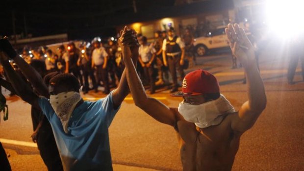 Protestors wearing masks raise their hands above their heads as they stand in front of police officers monitoring nighttime demonstrations in Ferguson.
