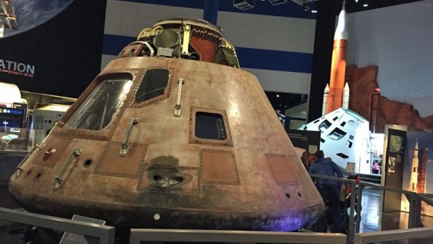 The actual Apollo 11 Command Module, battered  from reentry, with Orion and its SLS rocket behind.