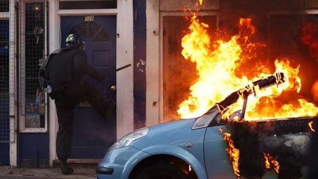 A riot police officer attempts to break down the door of a house next to a burning car in Clarence Road in Hackney.
