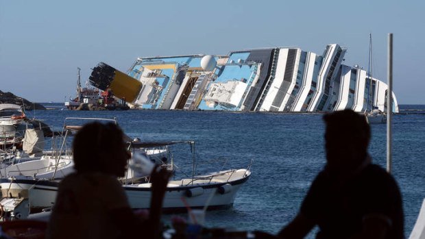 Restaurant patrons enjoy a drink while the wreck of the Costa Concordia looms in the background.
