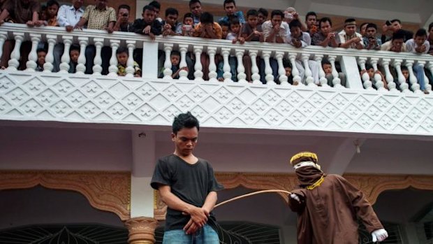 A masked and hooded official canes an Acehnese man for gambling.