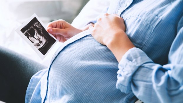 Pregnant woman keeping hand on belly and holding ultrasound image at home interiors. Pregnancy, parenthood, preparation and expectation concept. Close-up, copy space, indoors.