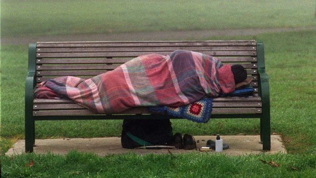 A homeless person lies bundled on a park bench.