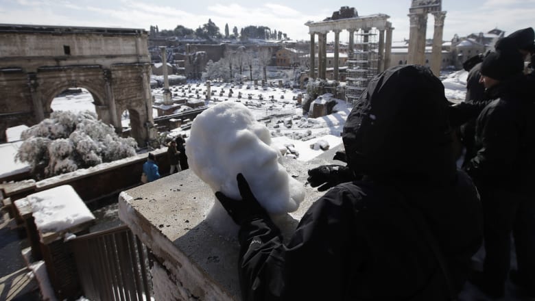 Sculptor Francesca Antonello moulds snow into a face as people look out towards the ancient Roman Forum covered in white.