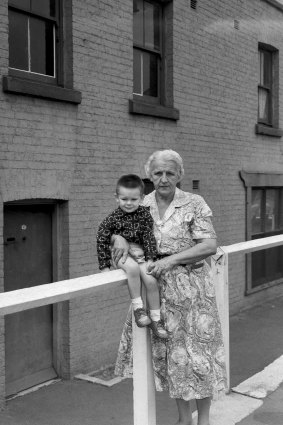 "MARGARET THOMPSON shows her two-year-old grandson PAUL the cottage in Susannah Place — dated 1844 — where his father was born. Mrs Thompson has lived around The Rocks all her life, and was baptised and married at St. Patrick's, Church Hill."