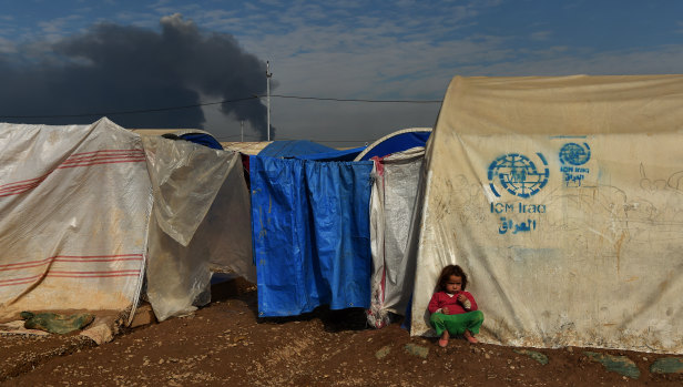 Smoke from the Qayyarah oil field fires fills the sky as a child sits in front of a tent in Qayyarah Airstrip Camp, home to over 13,000 displaced people.