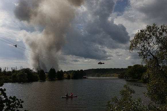People spend time near the river as a firefighting helicopter collects water to extinguish the fire that broke out in a distribution warehouse near Moscow after Ukraine’s drone attack.