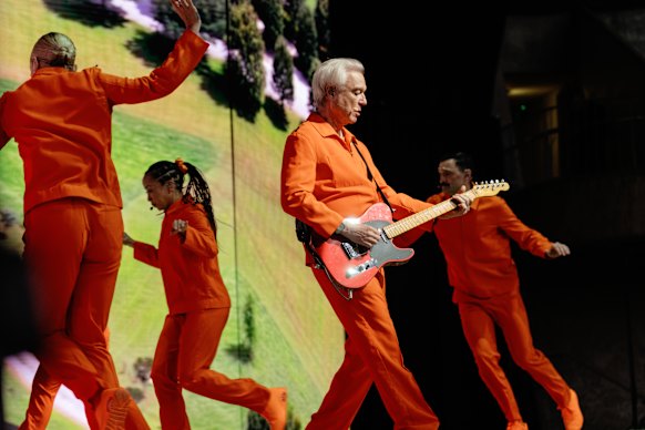 David Byrne performing at the Sidney Myer Music Bowl with his 12-strong crew of musicians and dancers, all dressed in orange boiler suits.
