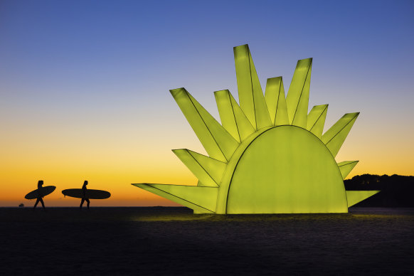 Surfers walk past a giant sun sculpture at sunrise on Mount Maunganui Beach, on the east coast of the North Island, New Zealand.