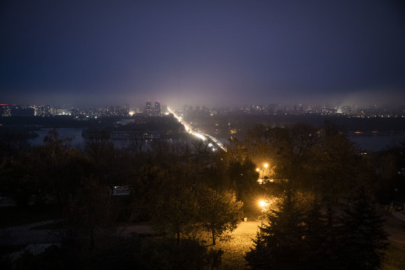 Cars cross the Metro Bridge and drive on the left bank between dark buildings at dusk in Kyiv, Ukraine. 