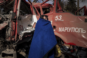 在一個一周沒有供暖的城市裡，一名婦女用毯子裹住自己，經過一輛被導彈擊中的馬里烏波爾消防車的外殼。 