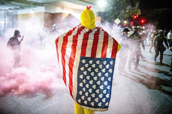 Un manifestant vêtu d'un costume de poulet et d'un drapeau américain avec des cœurs au lieu d'étoiles regarde les agents des forces de l'ordre tenter de disperser les manifestants près d'un centre américain d'immigration et de douane à Portland.