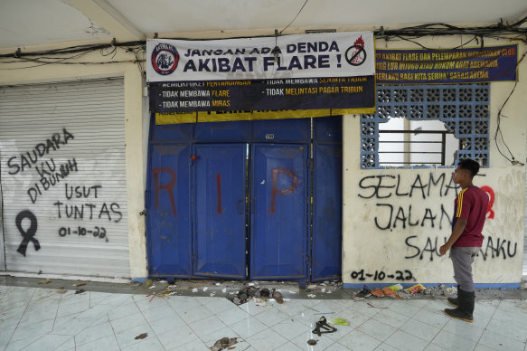 A man stands near a broken gate with graffiti “my brother was killed, investigate thoroughly,” and “Good bye my brother” at Kanjuruhan Stadium.